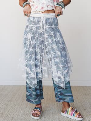 A layered, free-spirited look featuring a sheer floral lace skirt over wide-leg camo pants, styled with stacked boho bracelets and colorful platform sandals — the kind of outfit that doesn't follow rules and doesn't need to.