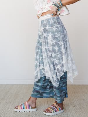 A sheer cream lace midi skirt with a handkerchief hem layered over blue camo leggings, styled with a floral top, stacked turquoise bracelets, and rainbow platform sandals — the kind of mix-and-match that feels totally intentional.
