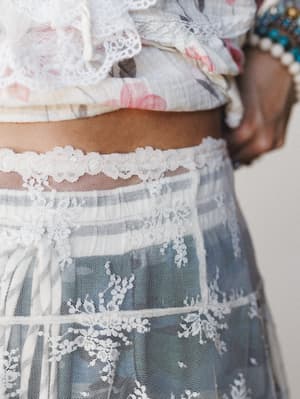 A close-up detail shot capturing the intricate lace overlay of a sheer cream skirt paired with a floral cropped top, with stacked bracelets just visible at the wrist — romantic and textural, like something you'd wear when the occasion calls for a lit