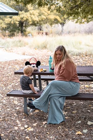 A woman wears the Relaxing Robin Wide Leg Pant - New Teal as she sits at an outdoor picnic table with a young child. A Mickey Mouse lunchbox, water bottle, and snacks are on the table amid trees and grass in the background.