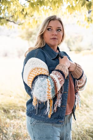 A woman stands outdoors under leafy branches, thoughtfully holding the collar of her Sukie Sweater Denim Jacket - Blue, which features chunky knit sleeves and fringe.