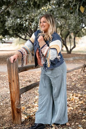 A woman with long blonde hair wears the Sukie Sweater Denim Jacket - Blue, featuring chunky knit sleeves and denim details, along with wide-legged pants and black shoes, as she smiles leaning on a wooden fence in a leafy woodland setting.
