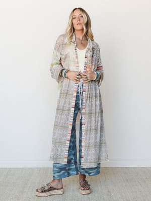 A long patchwork floral duster worn open over a cream textured tank and blue camo wide-leg pants, styled with stacked turquoise bracelets and platform sandals — the kind of outfit that looks effortless but clearly has a point of view.
