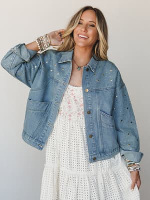 A smiling model layers a light-wash denim jacket covered in scattered gold star studs over a white tiered midi dress with floral and eyelet details — the kind of outfit that feels effortless and a little magical at the same time.