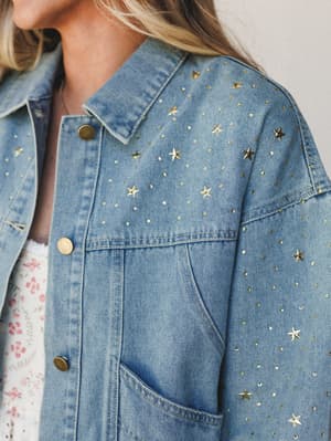 A close-up of a light wash denim jacket scattered with gold star studs and rhinestones, layered over a floral top — the kind of piece that makes a plain outfit feel like a whole moment.