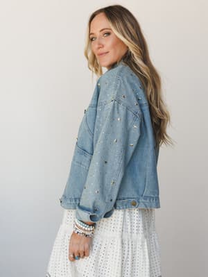 A star-studded denim jacket covered in gold star studs and rhinestones worn over a white eyelet tiered dress — the kind of piece that makes a white dress feel like a whole personality.