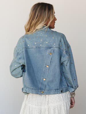 Shot from the back, this light-wash denim jacket is covered in gold star studs and rhinestone details — the kind of piece that makes a white eyelet skirt feel like a whole moment.
