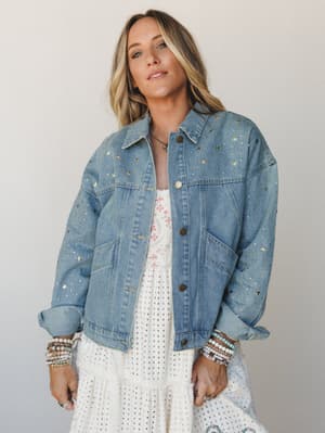An oversized light-wash denim jacket covered in gold and silver star studs, worn open over a white eyelet tiered dress with a floral-print bodice — the kind of outfit that looks like it was put together effortlessly but definitely wasn't.