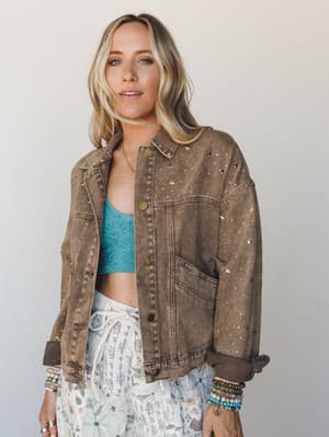 A washed mocha denim jacket covered in scattered gold and silver star studs is worn open over a teal crochet bralette and a white floral skirt, stacked with colorful bead bracelets — the kind of outfit that feels like it came together effortlessly bu