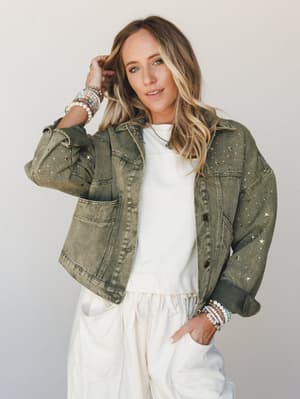 An olive acid-wash denim jacket scattered with gold star studs, layered over a cream sweatshirt and matching joggers — the kind of outfit that makes a Saturday errand run feel like a personality statement.
