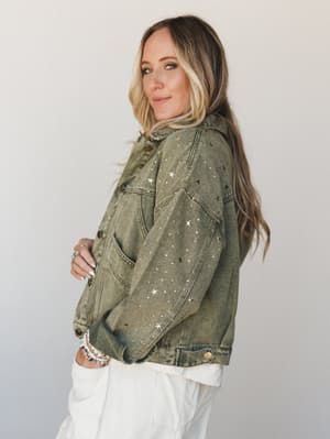 An olive acid-wash denim jacket covered in scattered gold and silver star studs, worn over white pants with stacked bracelets — the kind of jacket that makes a regular Tuesday feel like a little bit of a moment.