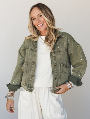 An olive green denim jacket covered in scattered gold star studs is layered over a cream top and wide-leg cream pants — the kind of outfit that feels effortless but clearly made choices.
