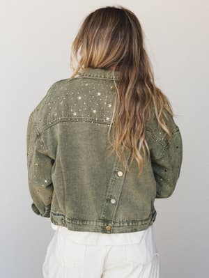A back-facing shot of an olive acid-wash denim jacket scattered with gold and silver star studs — the kind of jacket that makes a white outfit feel like a whole personality.
