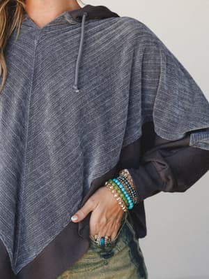 A close-up lifestyle shot of a cropped, asymmetric hoodie in heathered charcoal layered over a dark underlayer, styled with stacked beaded bracelets and a turquoise ring — that effortless mix of cozy and personal that makes an outfit feel like yours.