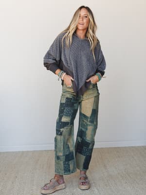 A relaxed full-body shot of a model in a charcoal oversized hoodie with an asymmetric hem paired with wide-leg patchwork jeans and platform espadrille sandals — the kind of outfit that looks effortless but is clearly very intentional.
