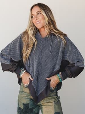 An oversized, draped hoodie top in heathered charcoal with contrasting dark panels and a relaxed poncho-like silhouette — styled with patchwork olive jeans and stacked turquoise bracelets for a lived-in boho feel.