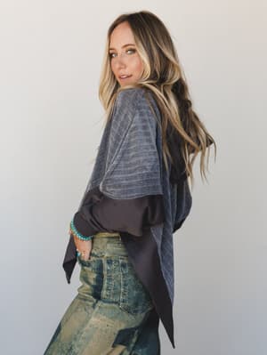 A model glances back over her shoulder in a slouchy two-tone hoodie top layered over acid-wash olive jeans, styled with turquoise bracelets — the whole look reads like a cool Saturday with nowhere to be.