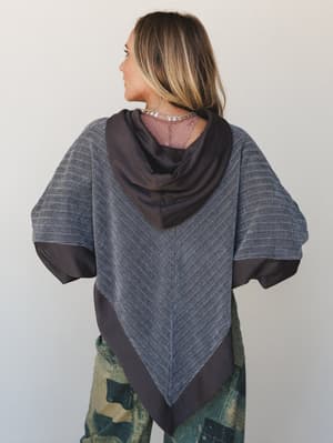A back-view shot of a mixed-fabric hoodie top in heathered charcoal and dark brown, styled with camo pants and a silver charm necklace — the asymmetric pointed hem and contrast hood give it a relaxed, artsy edge.