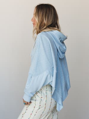 A relaxed side-profile shot of a mixed-fabric light blue hoodie with a dramatic asymmetric hem, paired with floral-embroidered white pants — the kind of effortless layers that feel intentional without trying too hard.