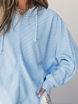 A soft, oversized light blue hoodie with a subtle diagonal texture, styled with a delicate chain necklace and a peek of floral bottoms — the kind of cozy you actually want to leave the house in.
