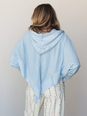 Shot from behind, a model pairs a light blue oversized hoodie — with a distinctive asymmetric pointed hem — over cream wide-leg pants covered in delicate embroidered floral vines, giving the whole look an easy, free-spirited feel.