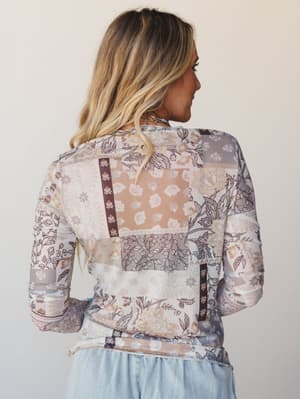 Shot from the back, a model in a long-sleeve mesh layering top covered in a patchwork-style mix of floral and paisley prints in warm mocha and dusty lavender tones, paired with light wash denim — the kind of top that makes a simple outfit feel intent