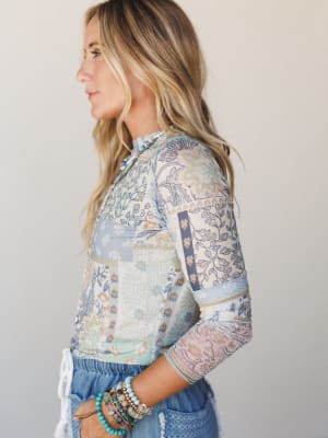 A side-profile shot of a model in a patchwork-print mesh layering top in dusty blue and cream, styled with denim shorts and stacked beaded bracelets — soft, expressive, and effortlessly put-together.