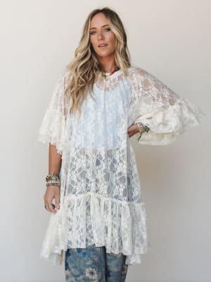 A flowy cream lace overlay dress with bell sleeves and a ruffled hem worn over a light blue slip and floral jeans, styled with layered beaded bracelets and colorful necklaces — effortlessly romantic and very much the kind of piece you'd emotionally a