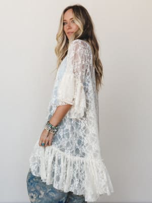 A long cream lace kimono-style layering piece worn over floral printed jeans, styled with stacked turquoise and beaded bracelets — romantic and free-spirited with a soft, dreamy quality.