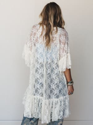 Shot from behind, a model wears a sheer cream lace longline kimono over a light blue top and floral jeans, the layered ruffled hem and stacked turquoise bracelets giving the whole look a relaxed, romantic boho feel.