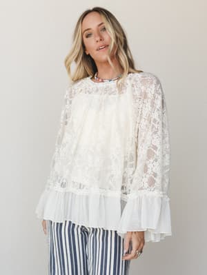 A dreamy cream lace top with sheer floral overlay and pleated ruffle hem, styled with navy stripe trousers and a colorful beaded necklace — romantic and a little unexpected.