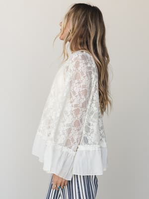 A cream lace top with sheer floral lace body and ruffle hem hem is shot in profile against a soft neutral studio backdrop, paired with navy striped wide-leg pants — romantic and effortlessly boho.