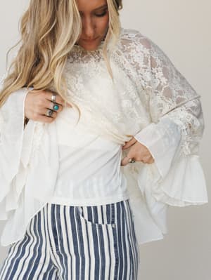 A dreamy cream lace top with dramatic bell sleeves and ruffle trim is styled over a white underlayer and tucked into navy striped wide-leg pants — the kind of outfit that feels like it found you at a vintage market and you never looked back.