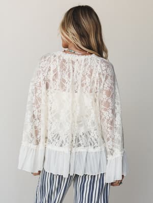 A sheer cream lace top photographed from the back, showing off its floral lace overlay, ruffle hem, and bell cuffs — styled with navy striped pants and layered bead necklaces for an effortlessly romantic, boho feel.