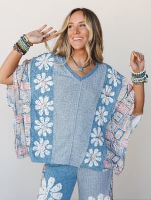 A smiling model poses playfully in a coordinating blue daisy-print set — an oversized ribbed poncho top and matching wide-leg pants — layered over a mixed patchwork kimono, stacked with colorful bracelets and a turquoise pendant for that effortlessly