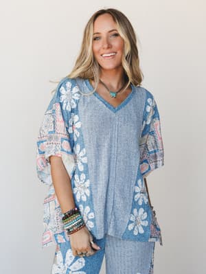 A smiling model in a relaxed, oversized mixed-media top — ribbed knit center panel meets floral daisy border and boho patchwork sleeves in soft blue and blush, styled with stacked stone bracelets and a turquoise pendant for that effortlessly collecte