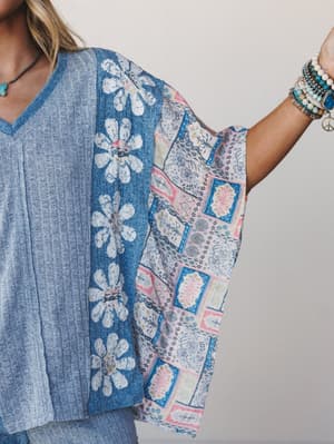 A close-up of a mixed-media top that layers heathered blue knit with a flowy patchwork floral sleeve panel — styled with stacked beaded bracelets and a turquoise pendant necklace for that effortless, collected boho feel.