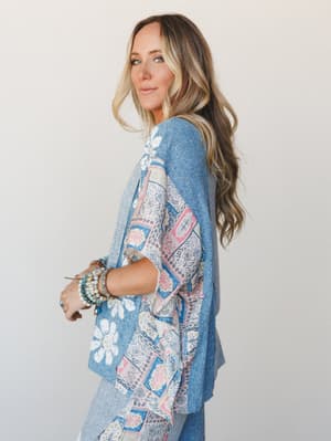 A relaxed mixed-media top in soft blue with a floral and patchwork print panel — styled with stacked beaded bracelets and an easy, over-the-shoulder glance that feels like a Saturday with nowhere to be.