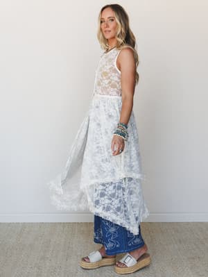 A flowy, sheer cream lace maxi dress layered over embroidered denim midi skirt, styled with stacked turquoise bracelets and white platform sandals — the kind of outfit that looks like it came together effortlessly but absolutely did not.