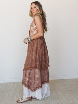 A mocha lace maxi dress with tiered ruffled skirt layers over a white underlayer, styled with stacked turquoise bracelets and Birkenstock-style sandals — the kind of dress that works just as hard at a wedding as it does wandering a farmers market.