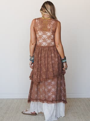 Shot from the back, a model wears a sheer mocha lace maxi dress layered over a white lace skirt, styled with stacked turquoise beaded bracelets and sandals — effortlessly boho with a soft, romantic feel.