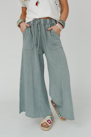Wearing the Relaxing Robin Wide Leg Pant in Faded Teal with a white short-sleeve top, colorful bracelets, and multicolored woven sandals, this boho-comfy look is finished with a casual pose and hands in pockets.
