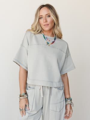 A relaxed matching set in soft dove grey — cropped sweatshirt top and drawstring pants styled with layers of colorful beaded necklaces and stacked stone bracelets, giving the whole look a lived-in, effortlessly personal feel.