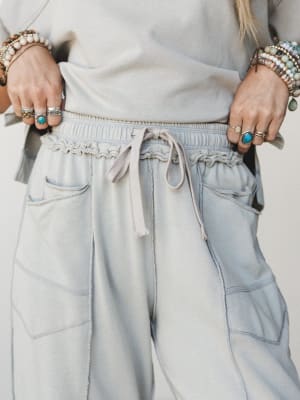 A close-up detail shot of soft dove grey wide-leg sweatpants with a ruched drawstring waistband, styled with stacked boho bracelets and silver rings — the kind of pants that feel like staying home but look like going somewhere.