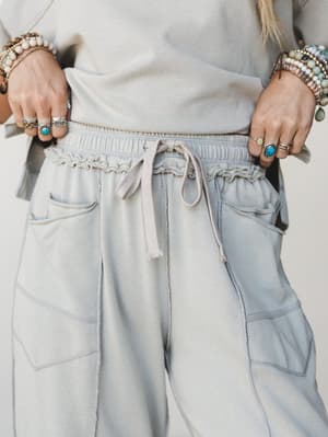 A close-up detail shot of soft dove grey wide-leg sweatpants with a ruched drawstring waistband, styled with stacked boho bracelets and silver rings — the kind of pants that feel like staying home but look like going somewhere.