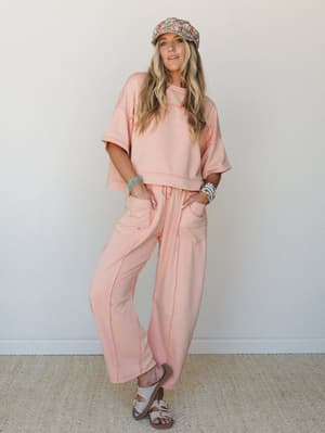 A matching dusty peach sweat set — cropped tee and wide-leg joggers with contrast stitching — styled with a floral newsboy cap, stacked turquoise bracelets, and Birkenstock sandals for an effortlessly relaxed, boho-casual feel.
