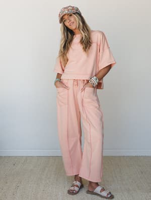 A matching peach-coral sweatshirt-and-wide-leg-pant set styled with a floral newsboy cap, beaded bracelets, and white buckle sandals — the kind of effortless outfit that looks like you tried just enough.