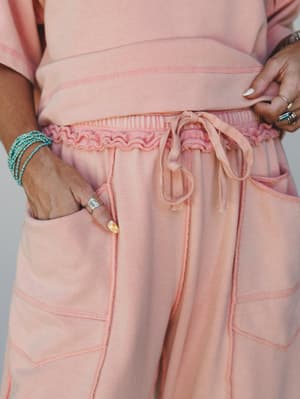 A close-up detail shot of a dusty pink matching set — the smocked, drawstring waistband and patch pockets of the wide-leg pants take center stage, styled with turquoise beaded bracelets and silver rings for that effortless boho finish.