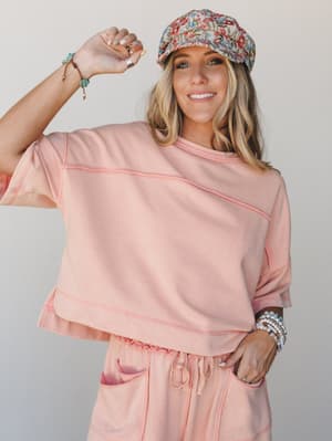 A breezy matching set in soft coral pink — cropped sweatshirt top and drawstring shorts styled with a floral newsboy cap and stacked bracelets, giving off that perfectly effortless 'ran an errand, looked great' energy.