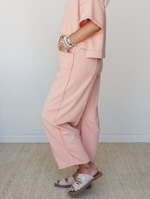 A side-profile shot of a matching peach-coral set — boxy crop top and relaxed wide-leg pants with contrast topstitching — styled with stacked beaded bracelets and double-buckle sandals for an easy, warm-weather feel.